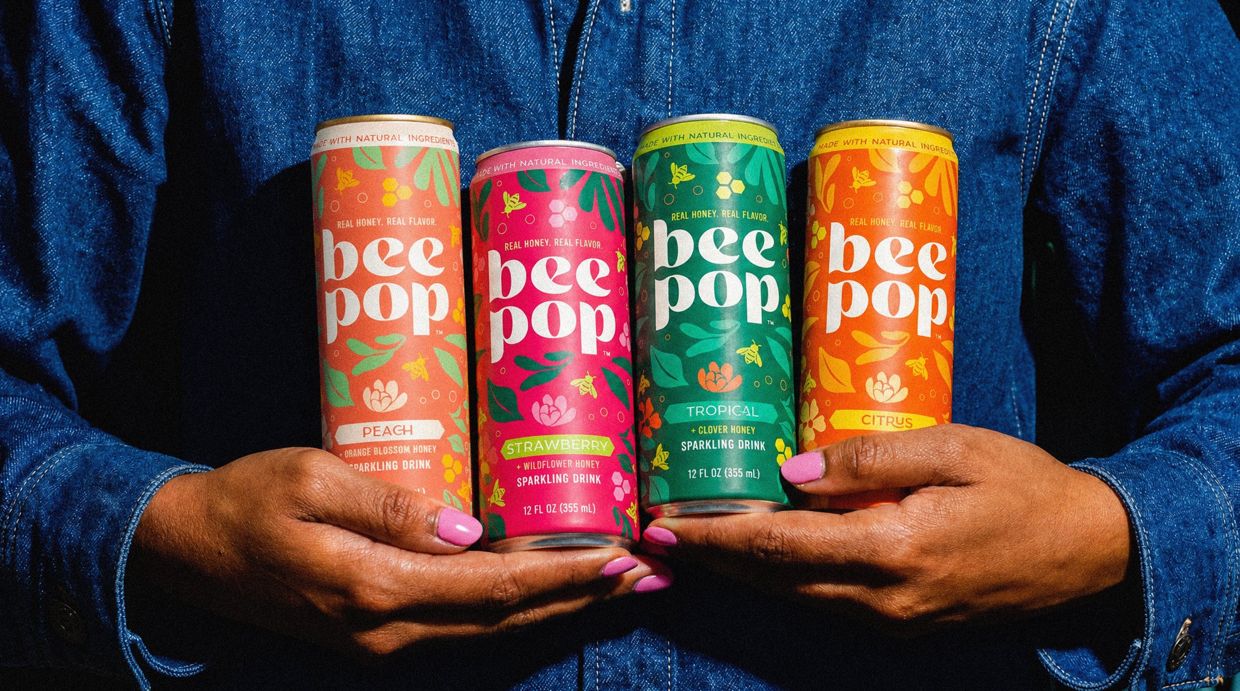 Four cans of 'bee pop' drinks held by a person wearing a blue denim jacket.