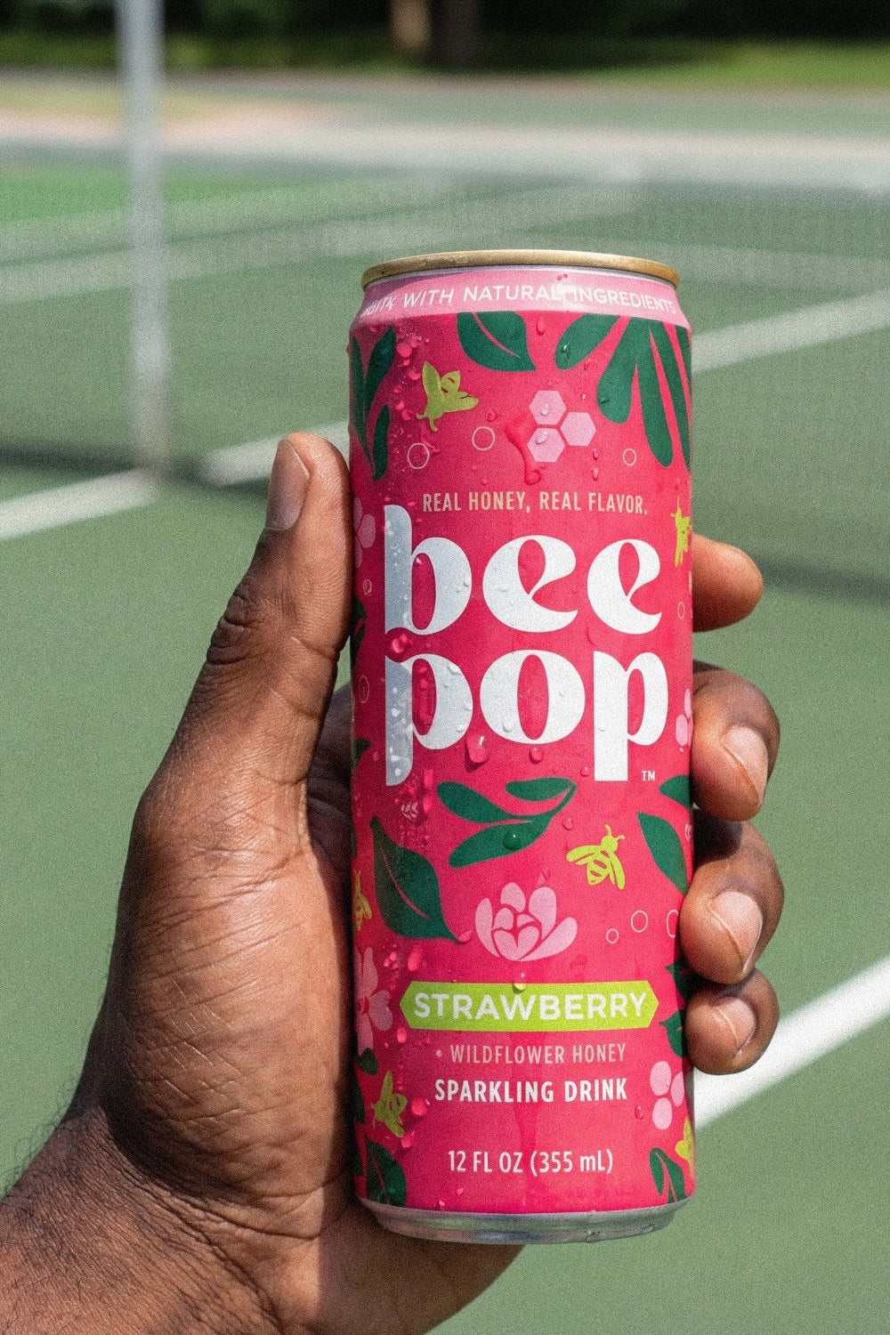 Hand holding a pink 'bee pop' can on a tennis court